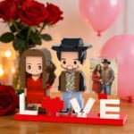 Custom brick figures of a couple with a “LOVE” base and heart detail, displayed with a reference photo, surrounded by roses and balloons in a romantic setting