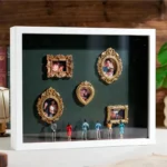 A white shadow box displaying several small golden-framed pictures of couples, family moments, and special occasions. Below the frames, miniature figurines of people are standing and admiring the photos. The background features soft lighting, with roses and pearls adding a decorative touch