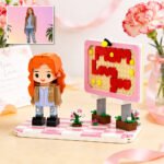 Custom brick figure of a girl with orange hair on a decorative base beside a signboard reading “Mom Love You,” with small flowers and a reference photo shown in the corner