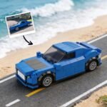 Custom blue car model made from building blocks placed on a road with a beach in the background. A photo of the real car version is shown above the model with an arrow pointing to it