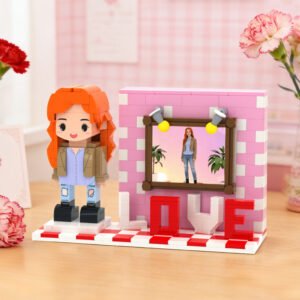 Custom brick figure of a girl with orange hair beside a pink display wall featuring a framed personalized photo and “LOVE” lettering, set on a decorative base with flowers in the background