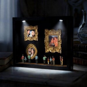 A personalized 3D figurine display with miniature people standing in front of framed photos of a couple. The images feature moments of love and affection, and the scene is lit with two small spotlights highlighting the frames