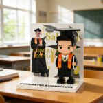 Custom brick figure of a male graduate in cap and gown holding a diploma, displayed on a base with “Congratulations I Love You” and a graduation photo backdrop in a classroom setting