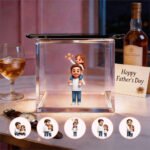 A personalized 3D holographic figurine of a father carrying his daughter on his shoulders inside a glass display box. Surrounding the display are smaller circular images showcasing different poses of the father and daughter. A 'Happy Father's Day' card sits next to a bottle of whiskey, with a glass of whiskey on the table, accompanied by a wristwatch