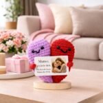 Crocheted heart-shaped couple in purple and red holding a personalized card with a mother and child photo and message, displayed on a table in a cozy living room setting