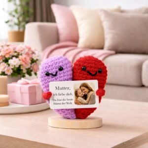 Crocheted heart-shaped couple in purple and red holding a personalized card with a mother and child photo and message, displayed on a table in a cozy living room setting