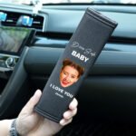 A personalized car seat belt cover featuring a woman's face with a wink, and the text 'Drive Safe Baby I Love You' along with the name 'James'. The cover is being held in a hand inside a car with a modern dashboard in the background