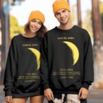 Couple wearing matching black sweatshirts with personalized moon phase design, names, date and romantic message, paired with orange beanies outdoors.