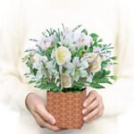 Person holding a small wicker basket filled with artificial white roses and greenery against a soft neutral background
