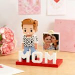 Custom brick figure of a mom standing on a base with “MOM” lettering, displayed with a photo of a mother and child, surrounded by flowers in a heartfelt gift setting