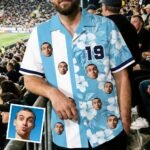 Person wearing a blue Hawaiian shirt with multiple cutout faces, in a stadium audience.